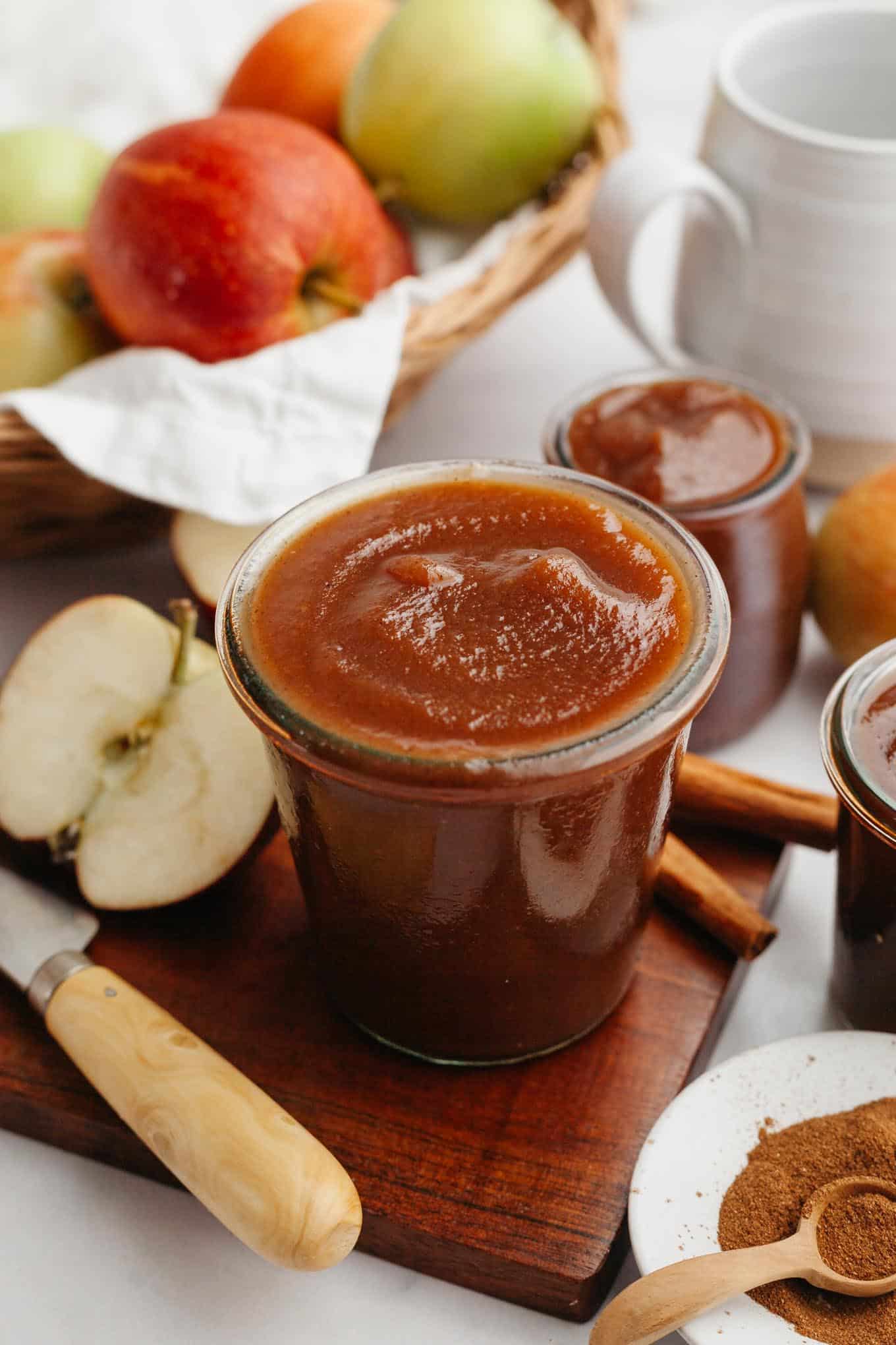 A jar of apple butter on a cutting board, with apples and spices surrounding it. 