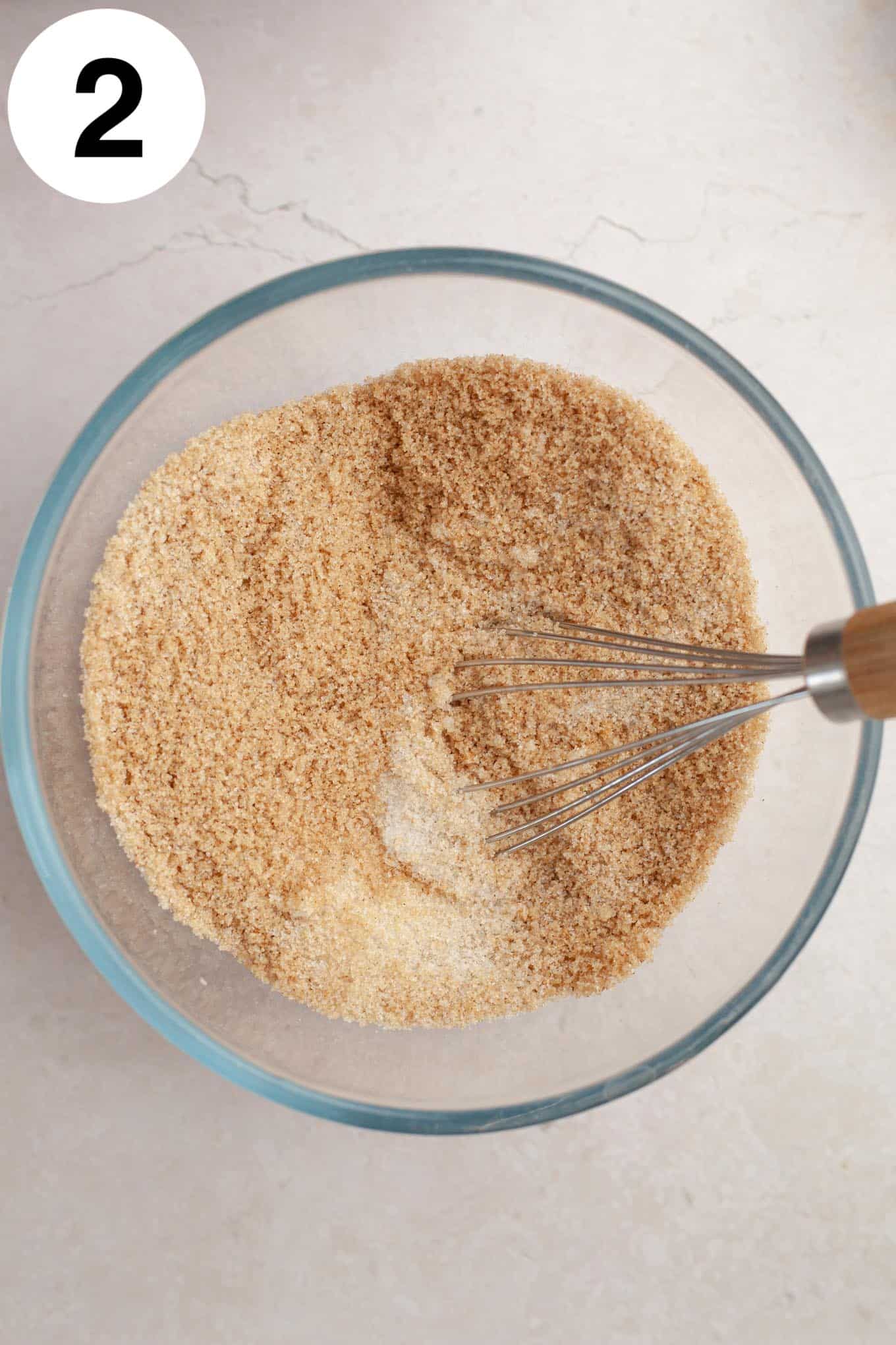 An overhead view of a sugar and spice mixture in a glass mixing bowl with a whisk. 