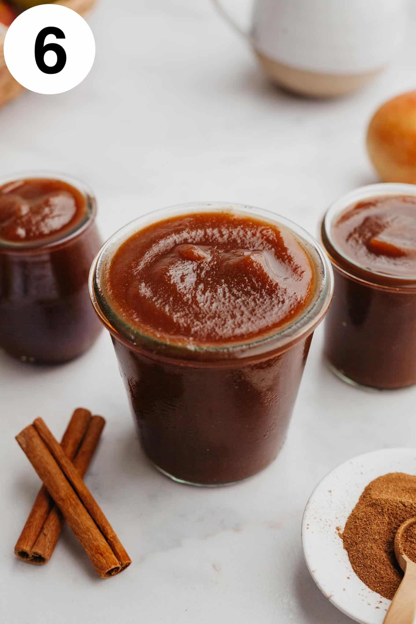 Three glass jars of apple butter, with cinnamon sticks and spices on the counter nearby. 