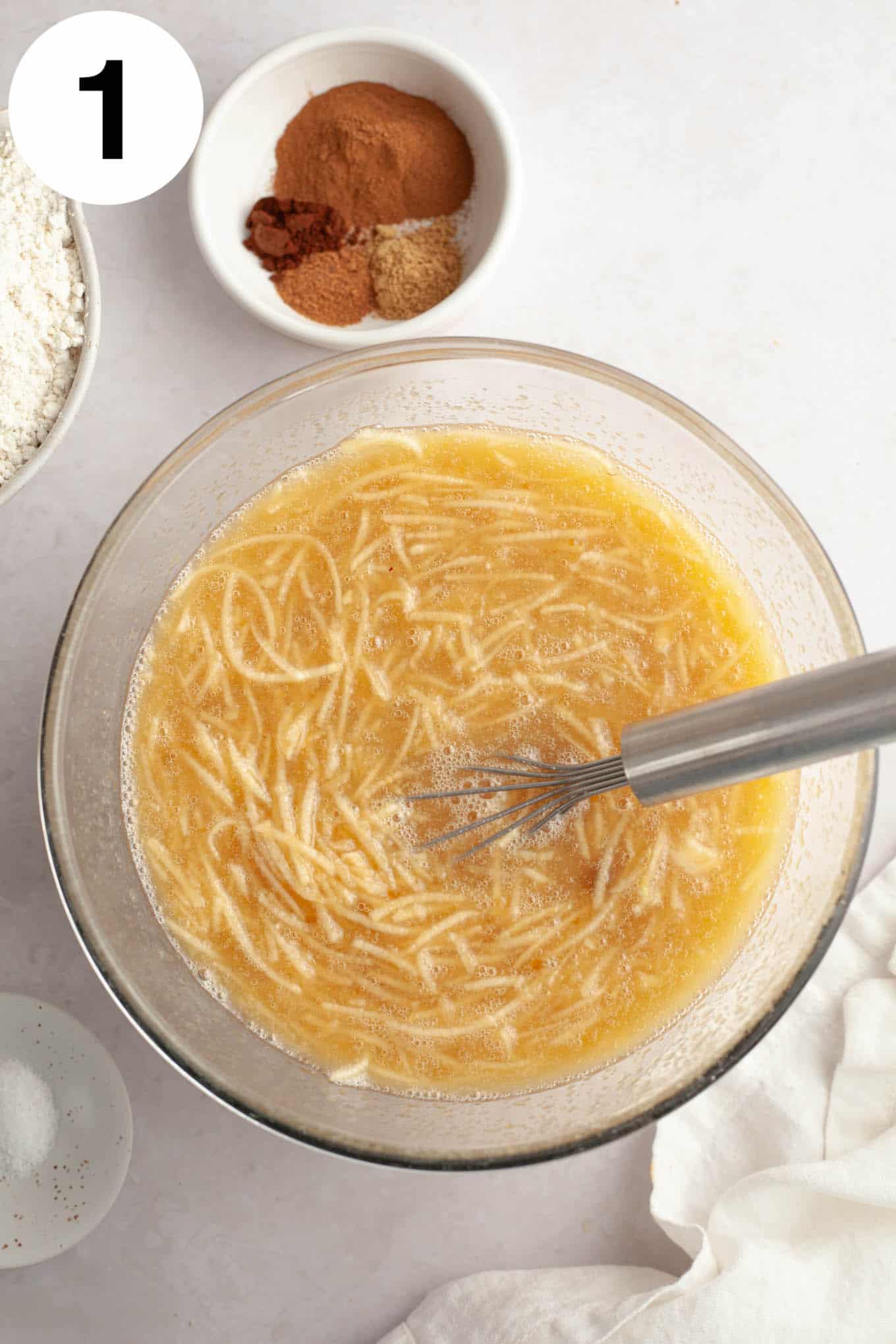 An overhead view of the wet ingredients with grated apple, in a glass mixing bowl with a whisk. 