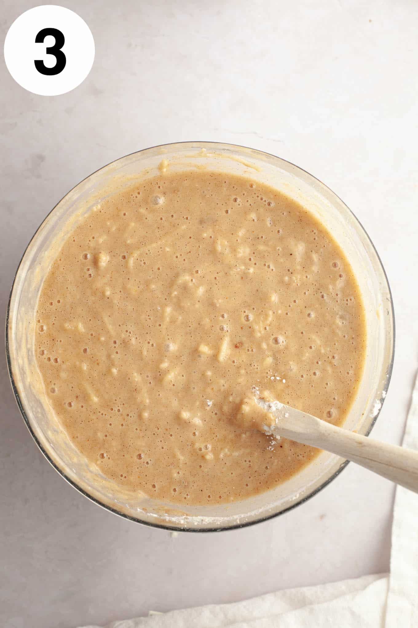 An overhead view of apple cake batter, in a glass mixing bowl with a wooden spoon. 