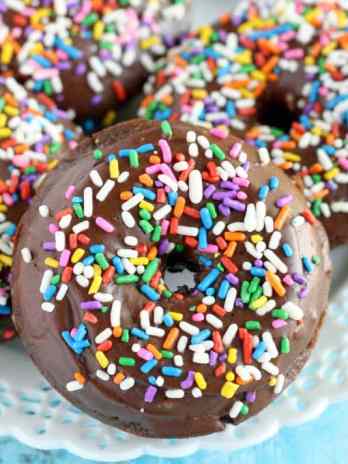Four baked chocolate donuts topped with chocolate glaze and sprinkles on a white plate.