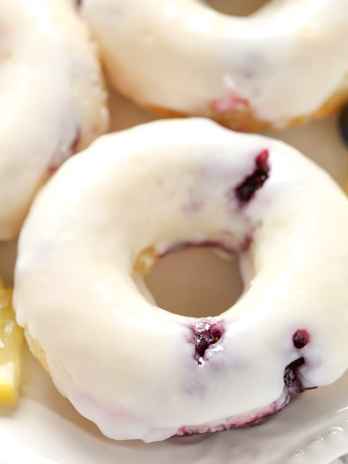 Overhead view of three lemon blueberry donuts on a white plate. Small lemon wedges and fresh blueberries surround the plate.