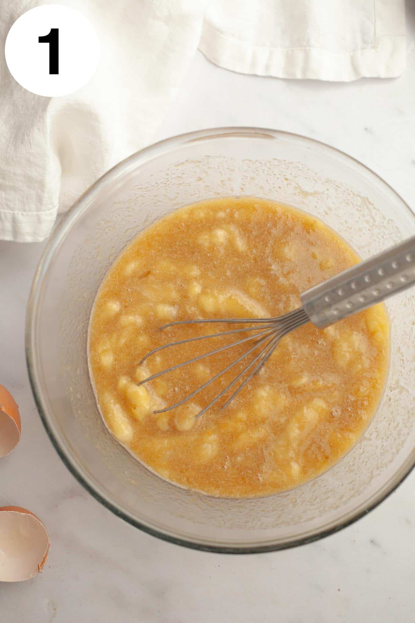 An overhead view of a banana and egg mixture in a glass mixing bowl, with a whisk. 