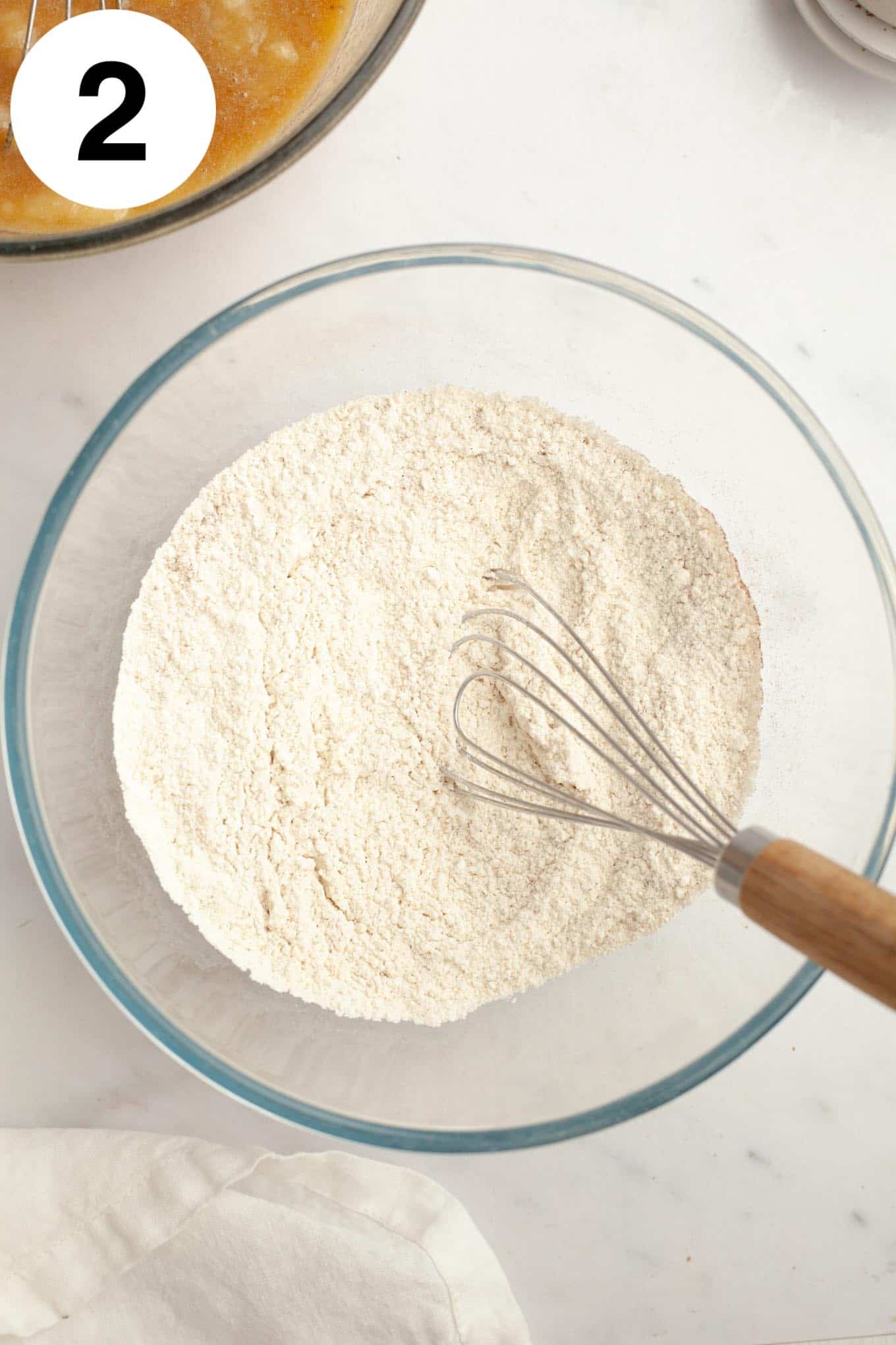 An overhead view of a flour mixture in a glass mixing bowl, with a whisk. 