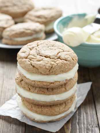 A stack of three carrot cake sandwich cookies on a square of parchment paper. A bowl of frosting and additional sandwich cookies rest in the background.