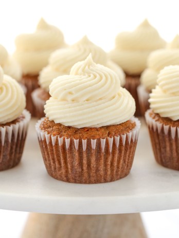A group of carrot cake cupcakes topped with cream cheese frosting setting on top of a marble cake stand.