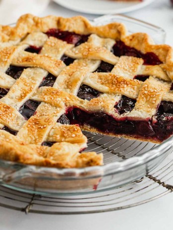 A cherry pie sitting on a wire rack with a slice taken out of it.