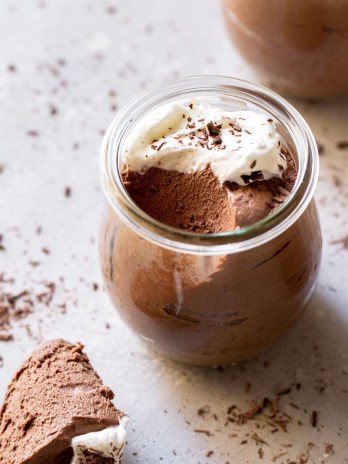 A glass jar filled with chocolate mousse topped with whipped cream and chocolate shaving with a spoonful taken out to show texture.