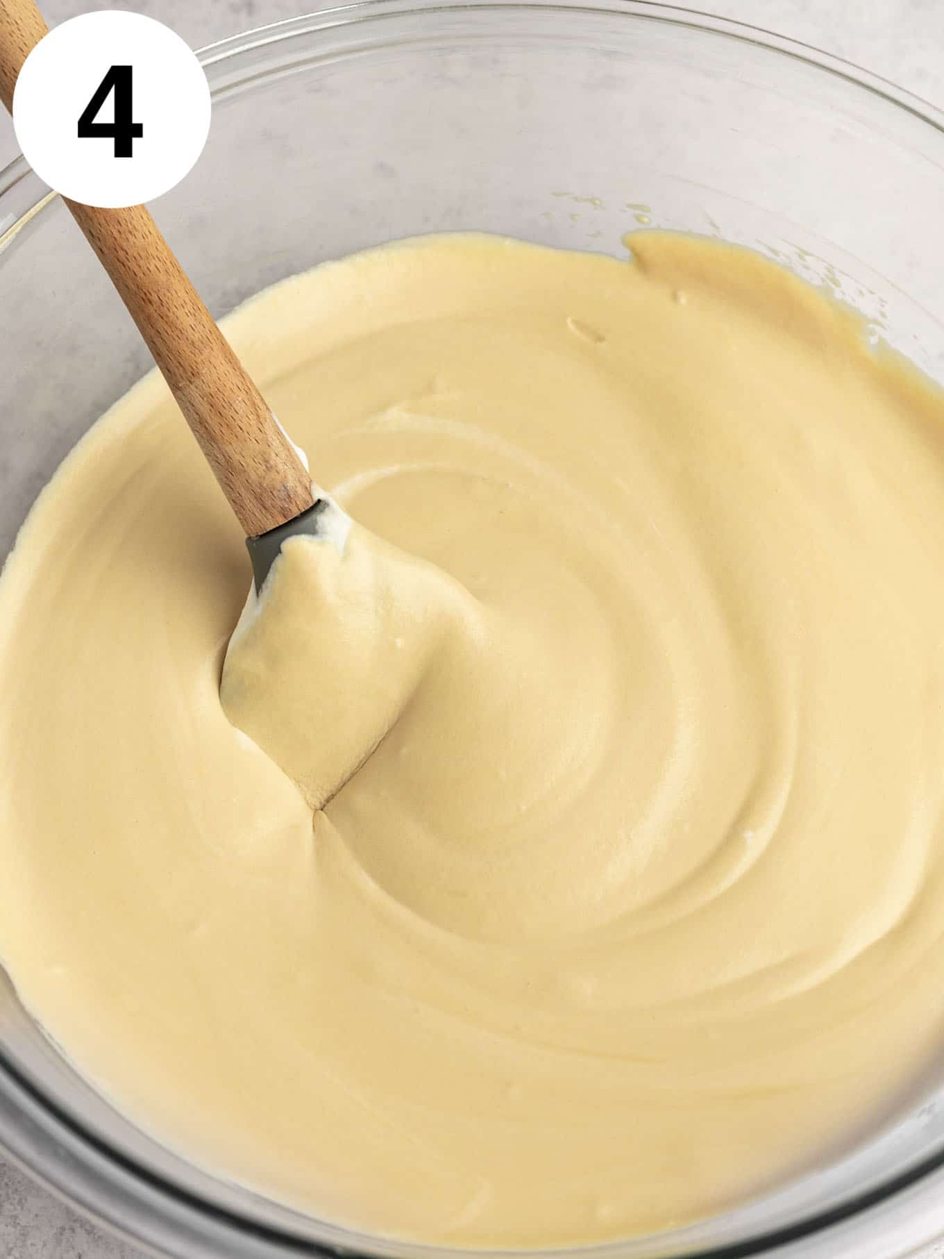 An overhead view of a coffee ice cream base in a glass mixing bowl, with a rubber spatula. 