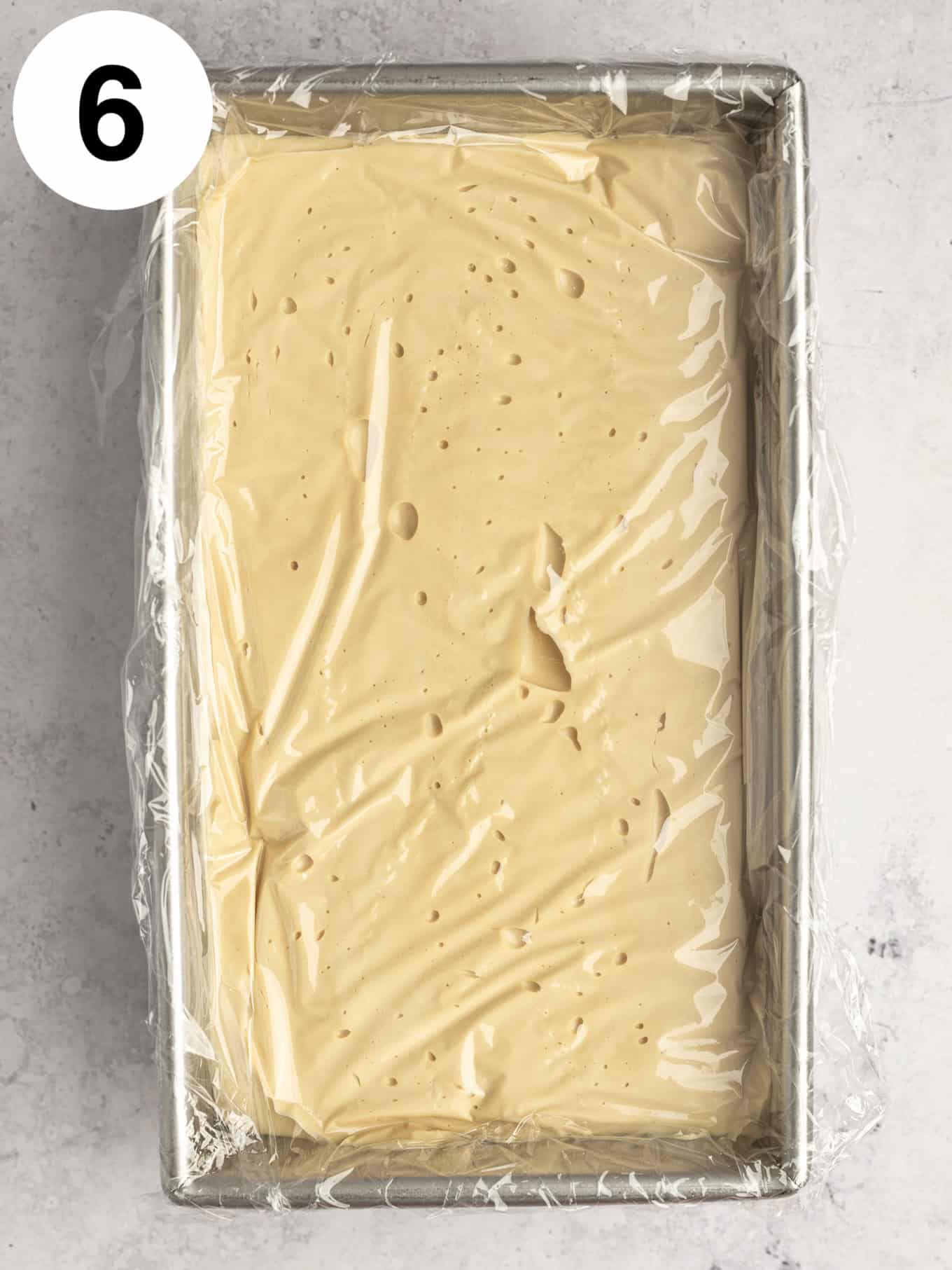 An overhead view of coffee flavored ice cream in a loaf pan, covered with plastic wrap. 