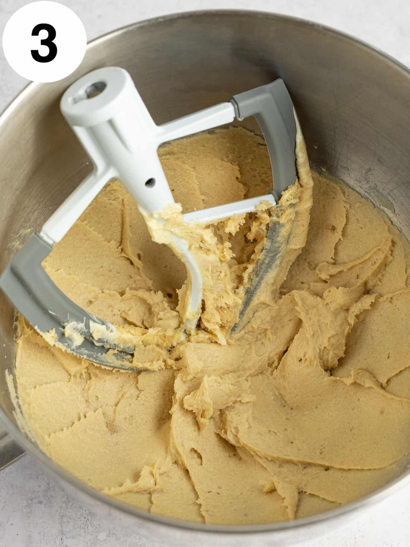 An overhead view of a beaten brown sugar-butter mixture in the bowl of a stand mixer, with a paddle attachment. 