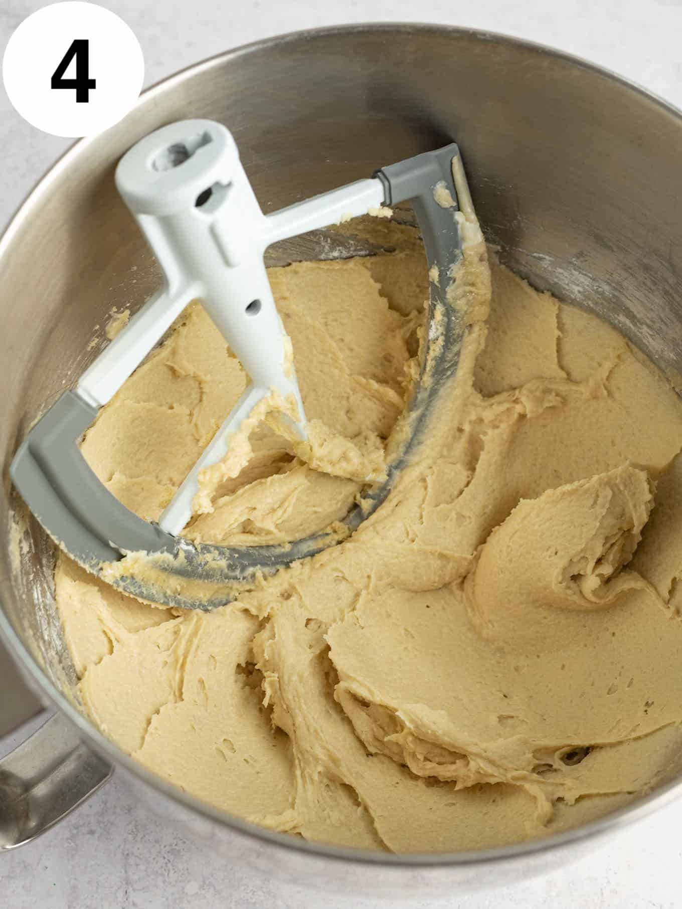 An overhead view of a beaten sugar-butter mixture in the bowl of a stand mixer, with a paddle attachment. 