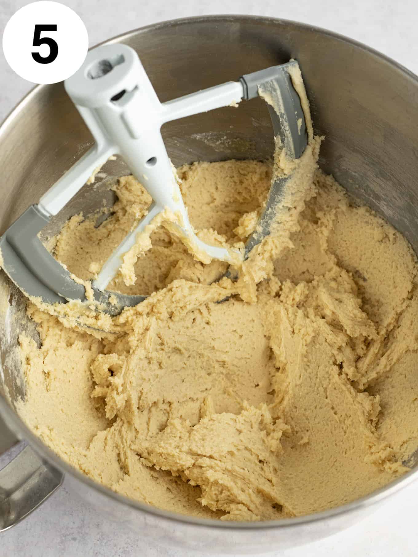 An overhead view of cookie dough frosting in the bowl of a stand mixer, with a paddle attachment. 