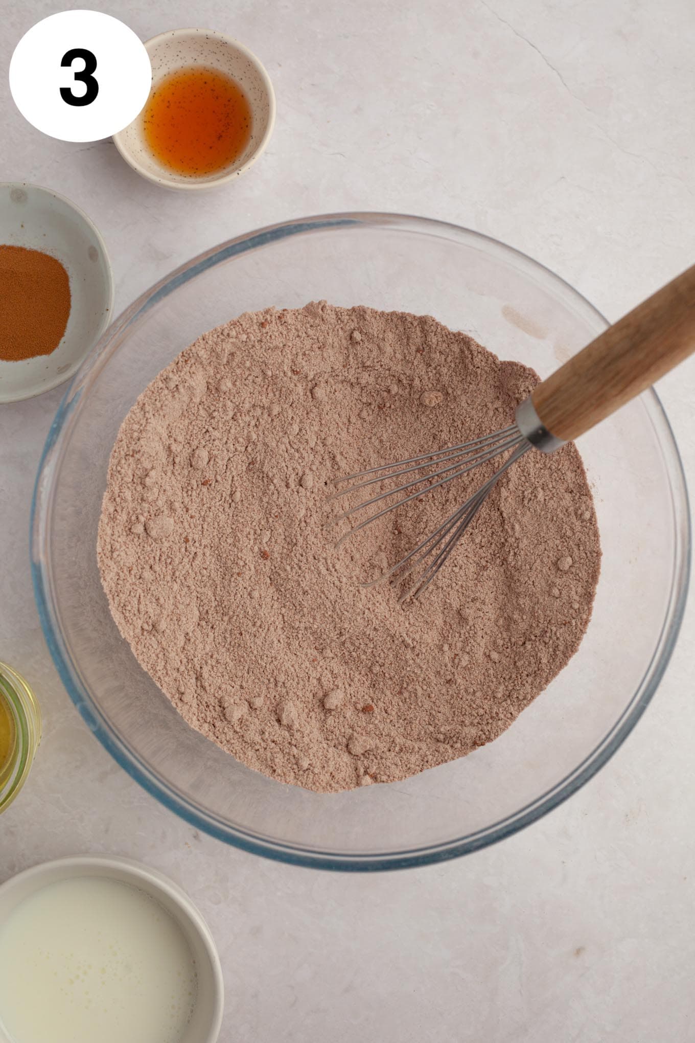 An overhead view of the dry ingredients in a glass mixing bowl, with a whisk. 