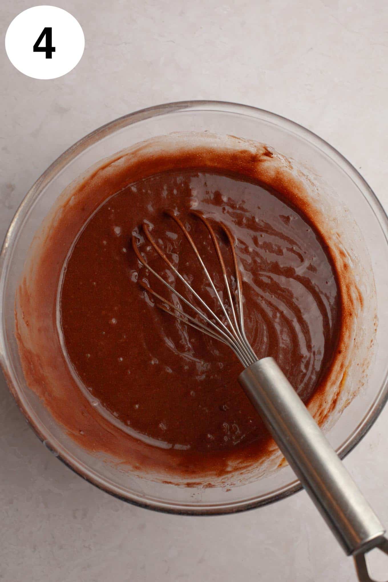 An overhead view of the wet ingredients for chocolate cupcakes in a glass mixing bowl, with a whisk. 