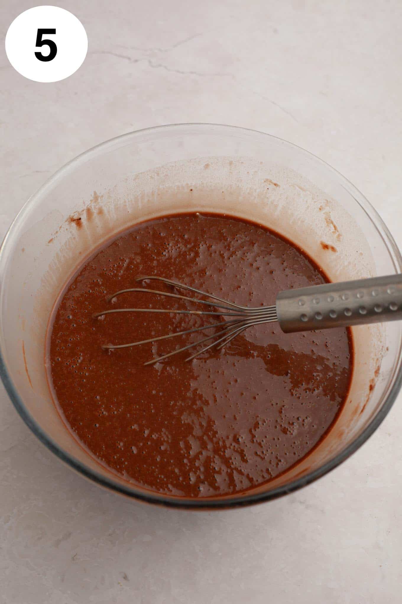 An overhead view of chocolate cupcake batter in a glass mixing bowl, with a whisk. 