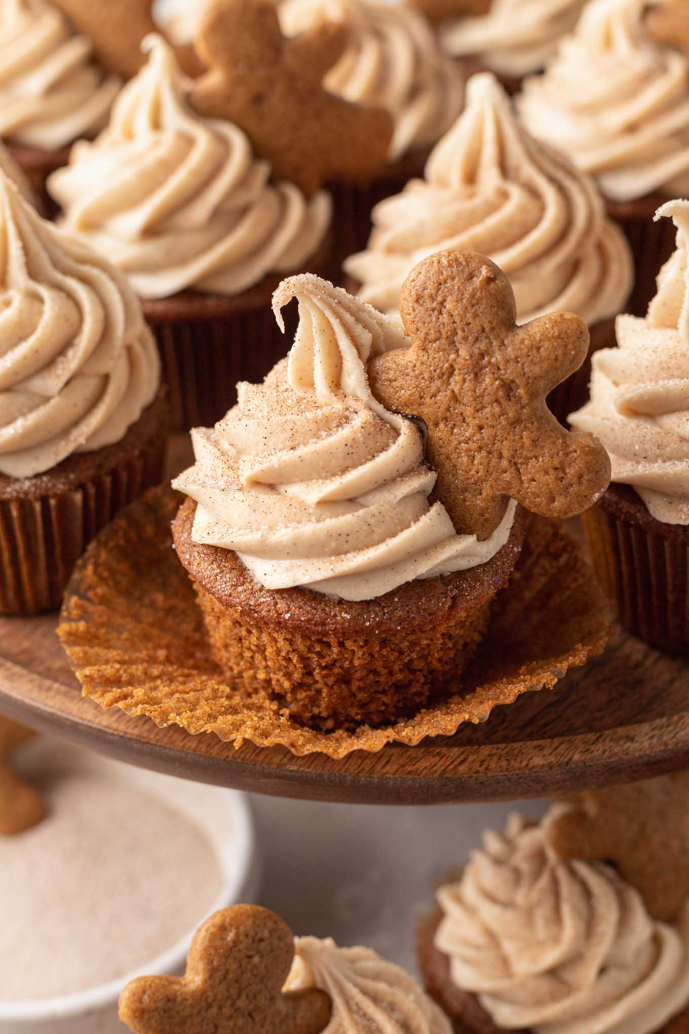 A gingerbread cupcake with the cupcake liner pulled down. More cupcakes are behind it on a cake stand.