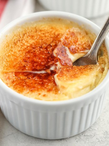 A close-up picture of a white ramekin filled with crème brûlée and a spoon inside of it.