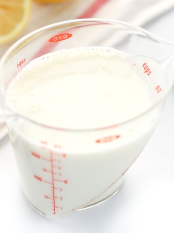 A clear measuring cup filled with homemade buttermilk along with a white napkin and lemons in the background.