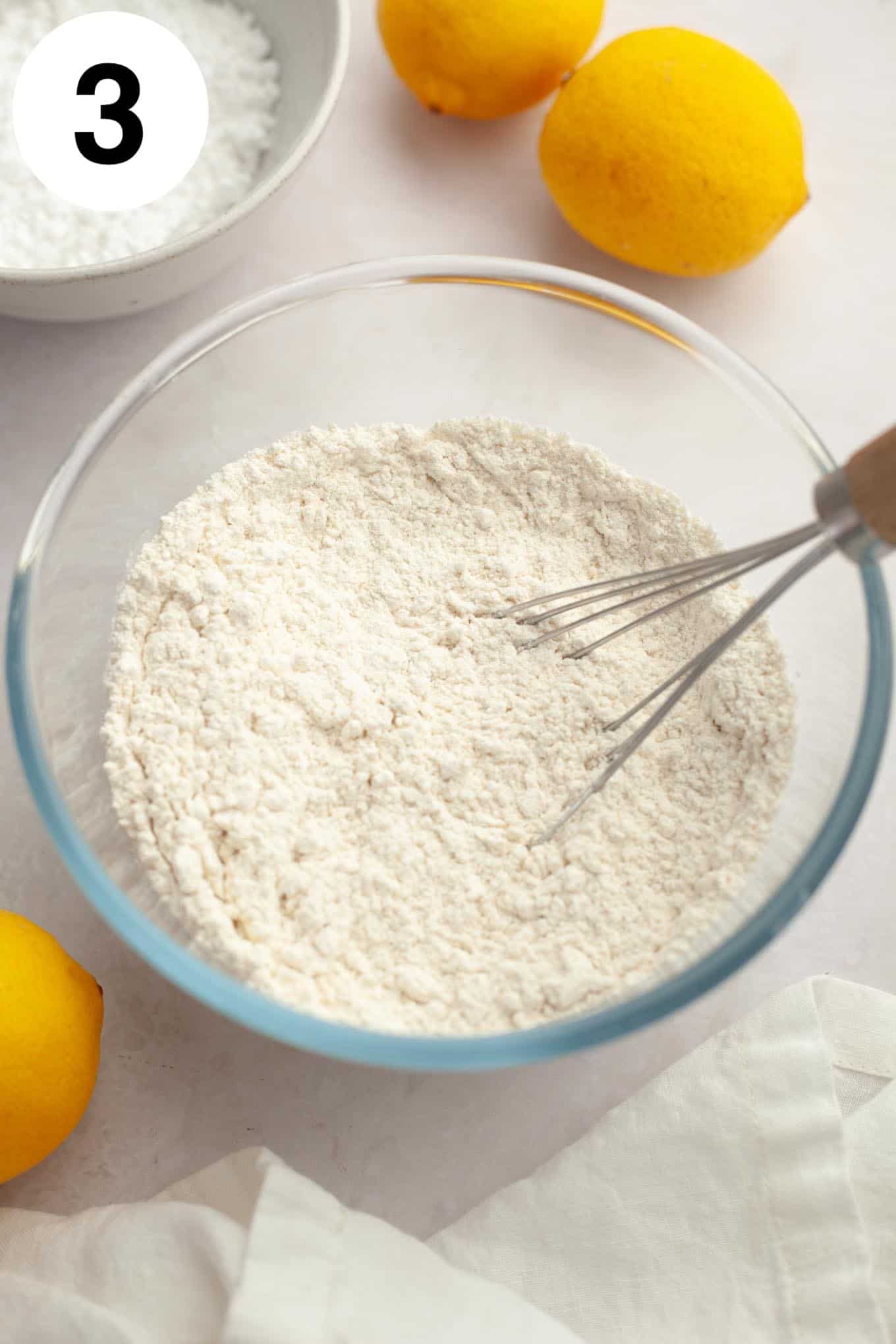 An overhead view of flour, salt, and baking powder in a glass mixing bowl with a whisk.  