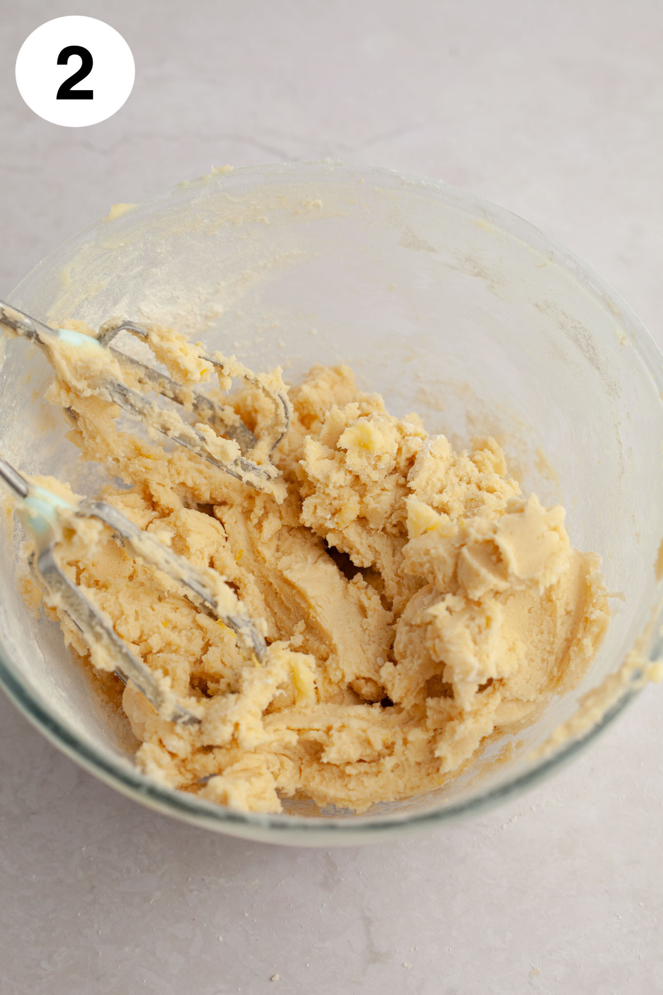 An overhead view of lemon cookie dough in a glass mixing bowl, with electric beaters.