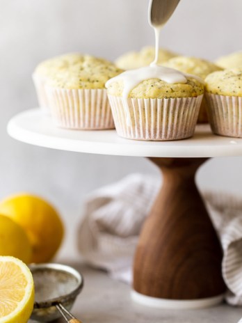 A marble cake stand with lemon poppy seed muffins being topped with a lemon glaze.
