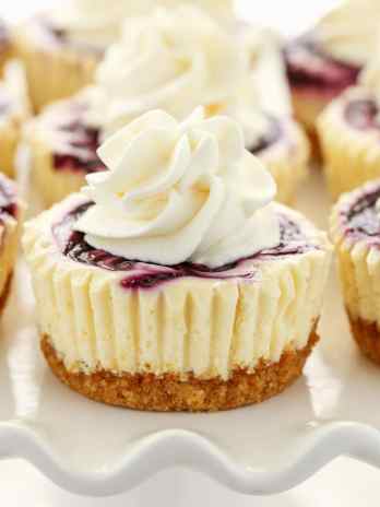Side view of lemon blueberry cheesecake bites topped with whipped cream on a white cake stand.