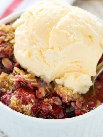 A white ramekin filled with mixed berry crisp with frozen berries, topped with a scoop of vanilla ice cream.