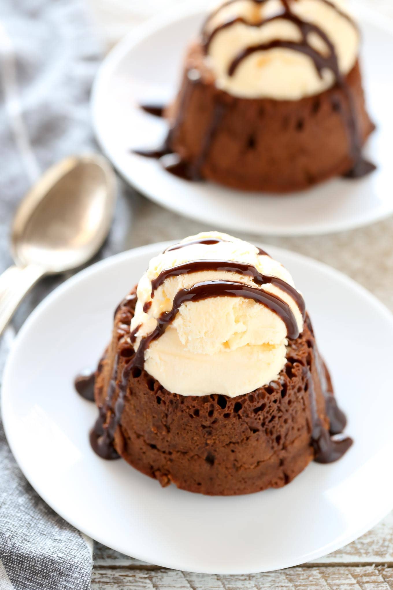 Two chocolate lava cakes topped with a scoop of ice cream and a drizzle of chocolate sauce. Both are on white dessert plates.