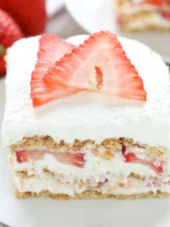 Close up of a slice of strawberry cream cheese icebox cake on a white dessert plate. Another slice and some fresh berries rest in the background.