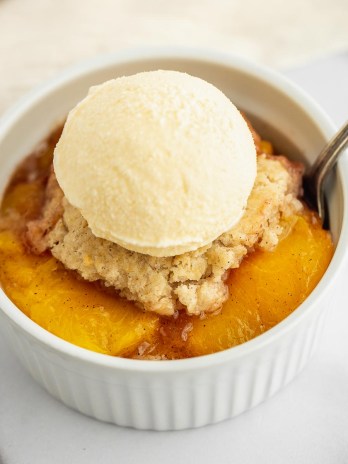 A white ramekin on a marble surface filled with peach cobbler and a scoop of ice cream on top.