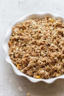 A round white baking dish with sliced peaches topped with the oat topping ready to be baked.
