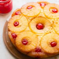 A whole pineapple upside down cake on a plate with a jar of cherries in the background.