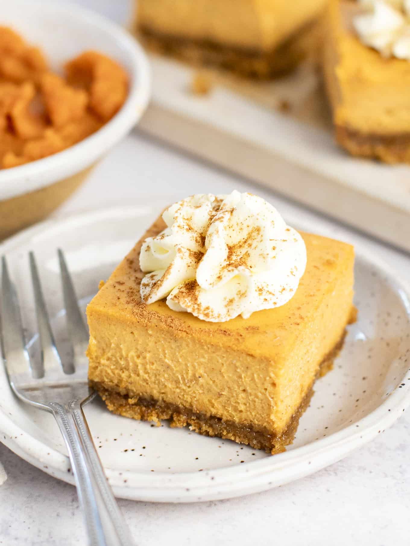 A close-up view of a pumpkin cheesecake bar on a dessert plate, garnished with whipped cream and pumpkin pie spice. 