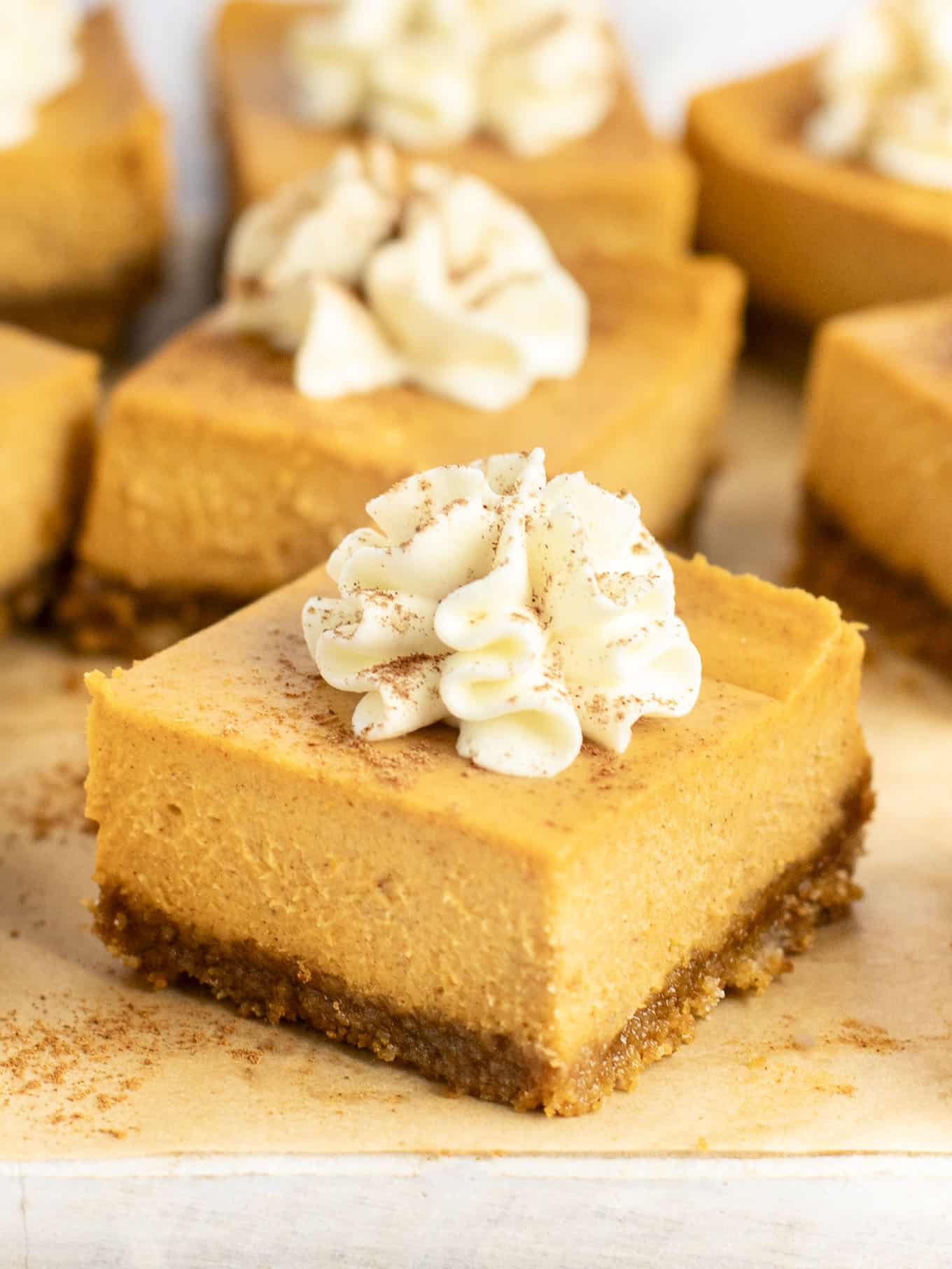 A close-up view of a pumpkin cheesecake bar garnished with whipped cream and pumpkin pie spice. 