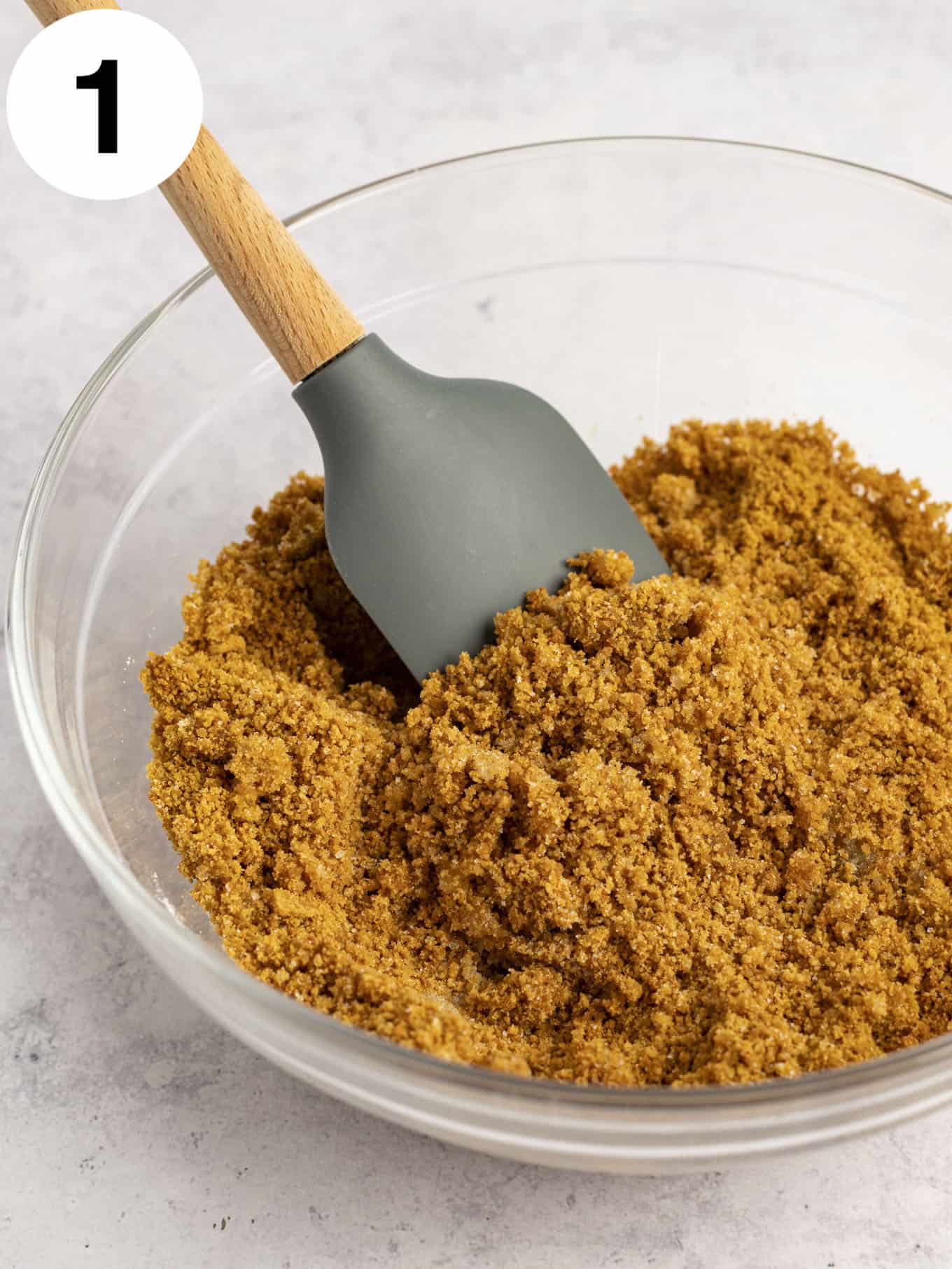 A gingersnap cookie crumb mixture in a glass mixing bowl with a rubber spatula. 