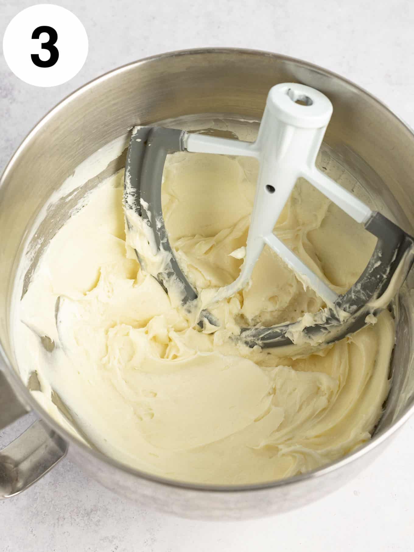 An overhead view of beaten cream cheese in the bowl of a stand mixer, with a paddle attachment. 