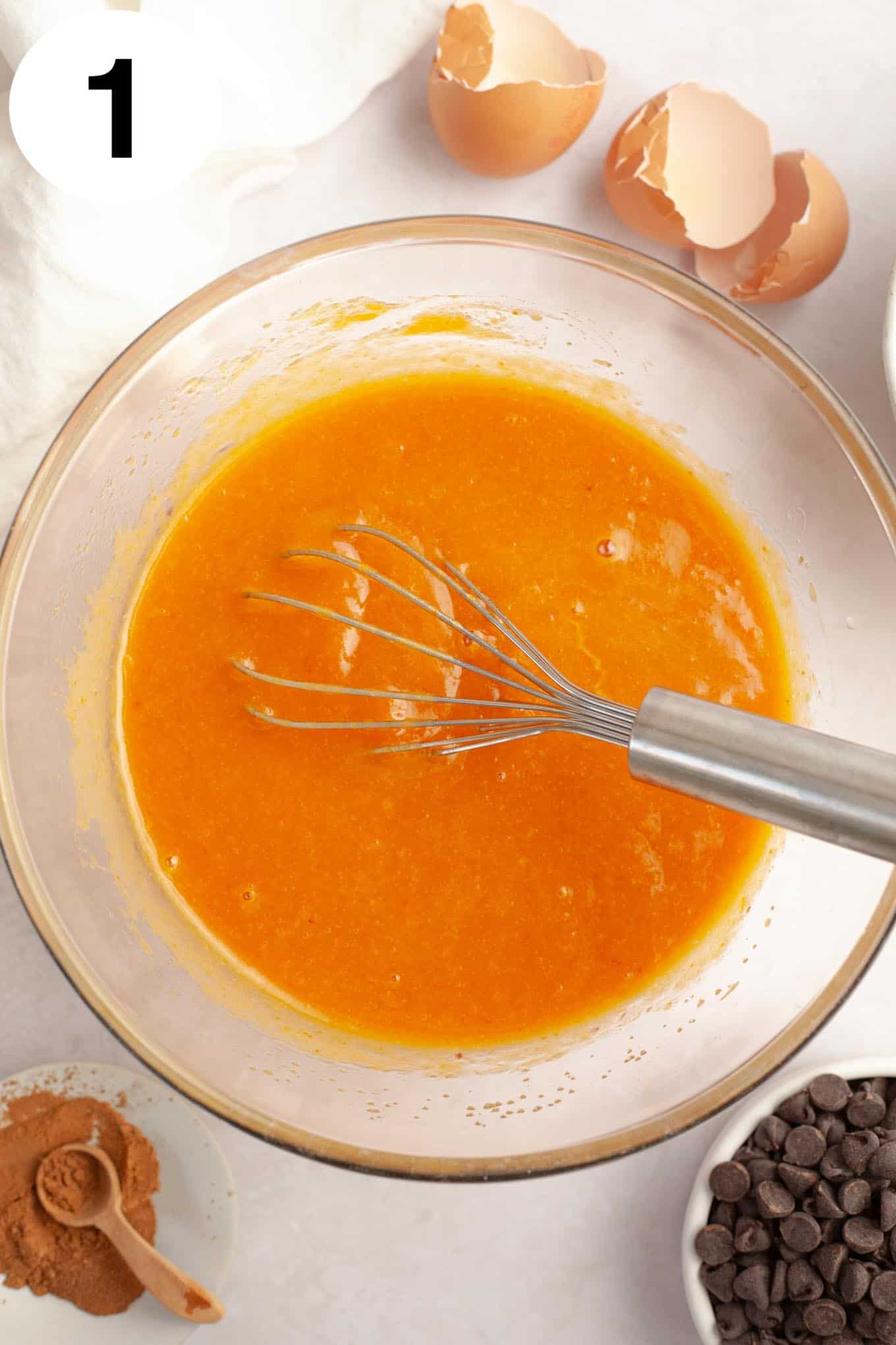 An overhead view of a wet pumpkin mixture in a mixing bowl, with a whisk. 