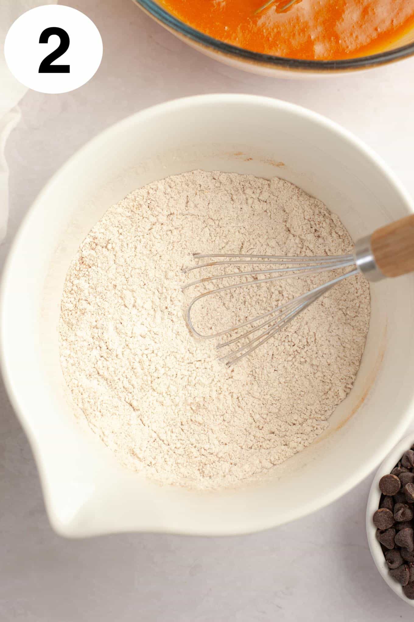 An overhead view of a dry flour mixture in a mixing bowl, with a whisk. 
