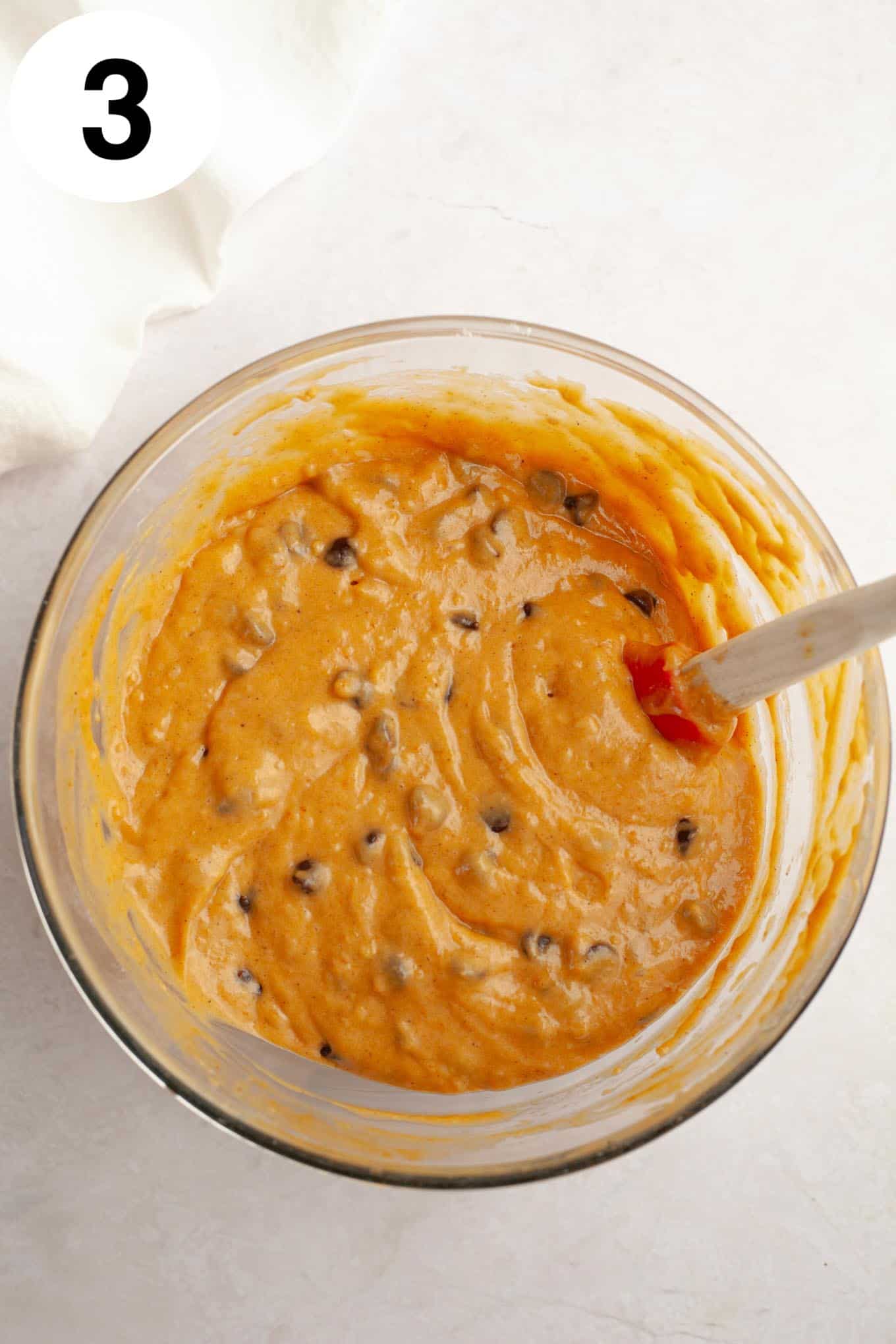 An overhead view of pumpkin chocolate chip muffin batter in a mixing bowl, with a rubber spatula. 