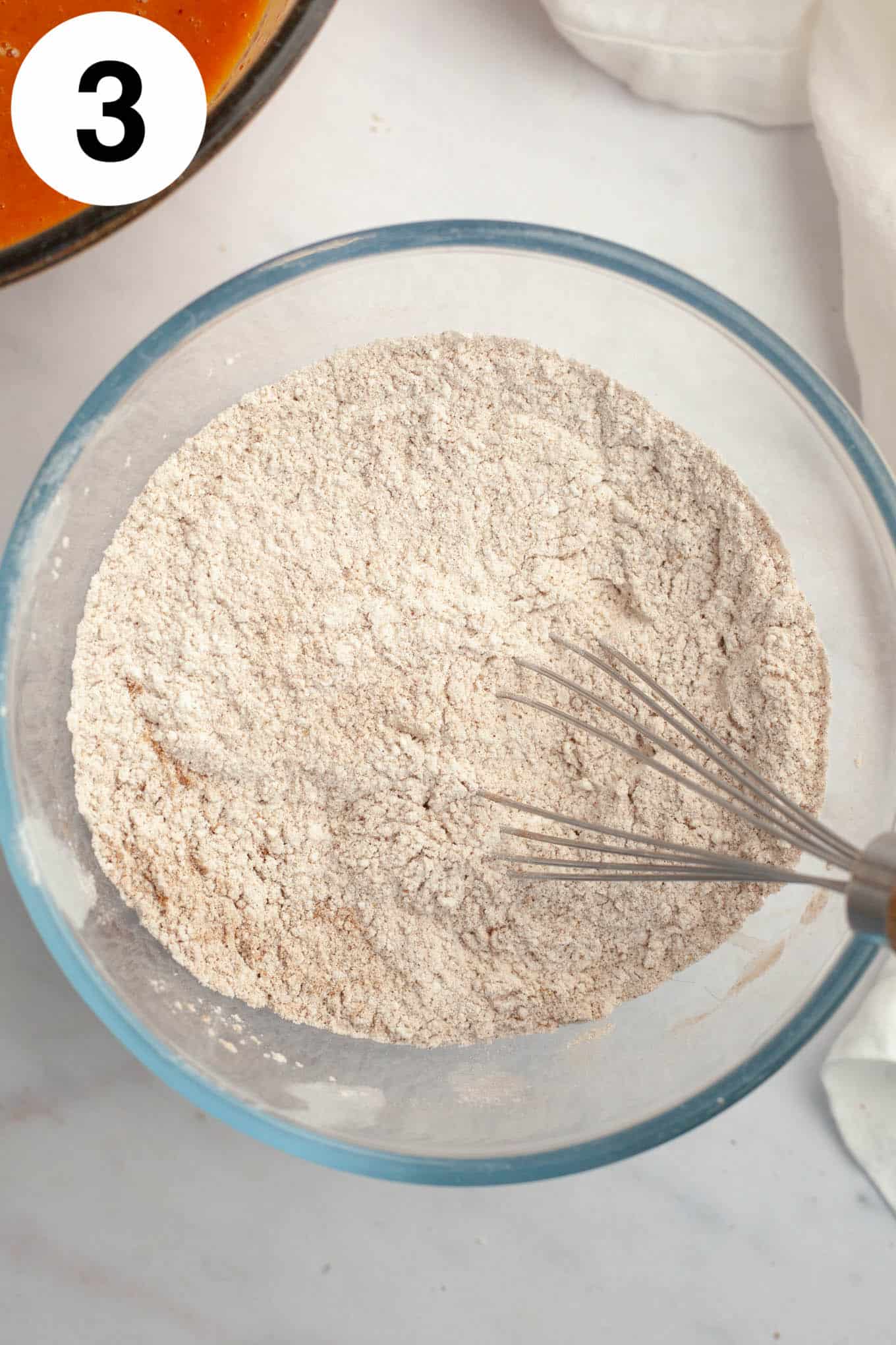 An overhead view of a dry flour mixture in a glass mixing bowl, with a whisk. 
