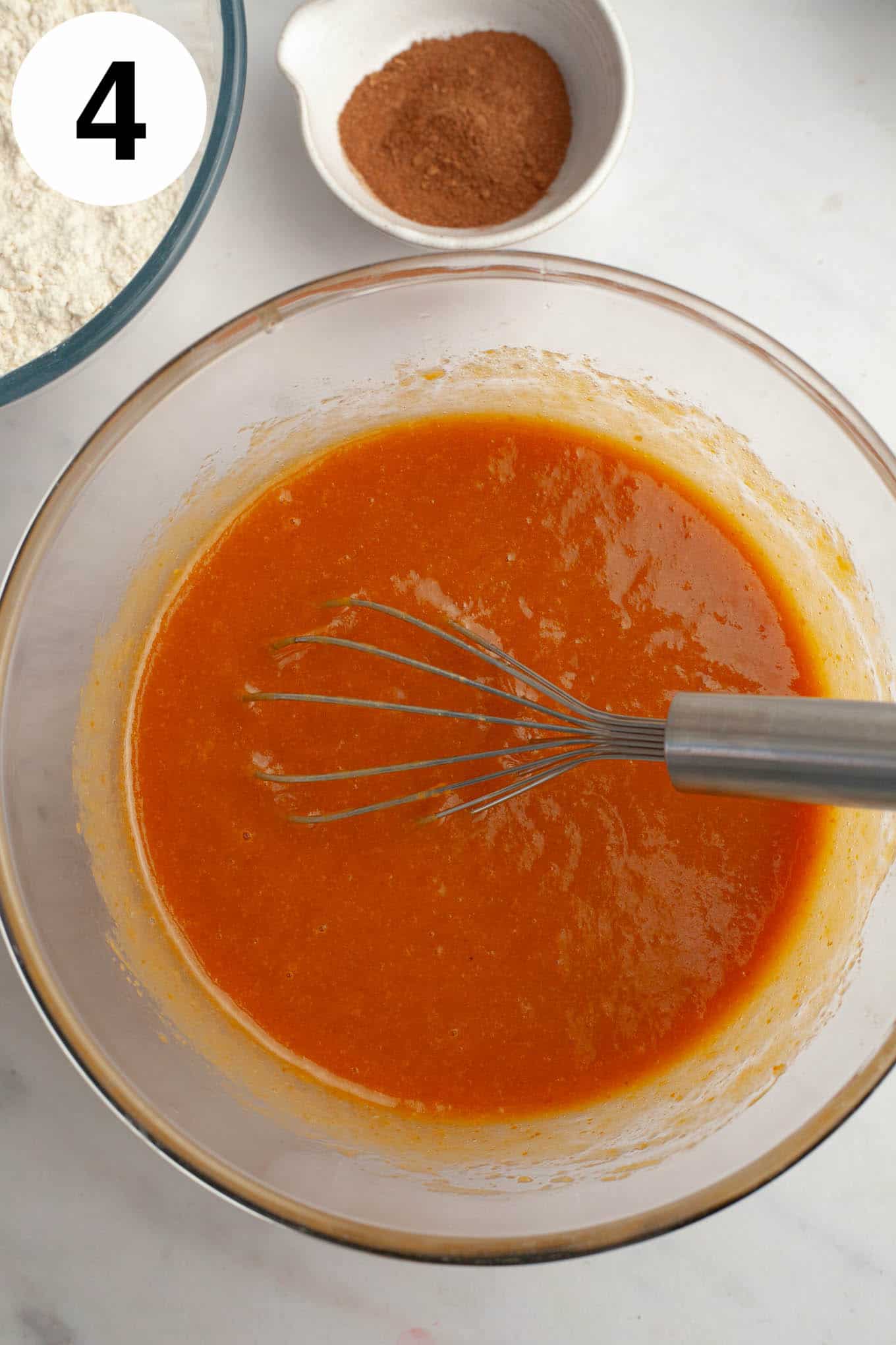 An overhead view of a wet pumpkin mixture in a glass mixing bowl, with a whisk. 