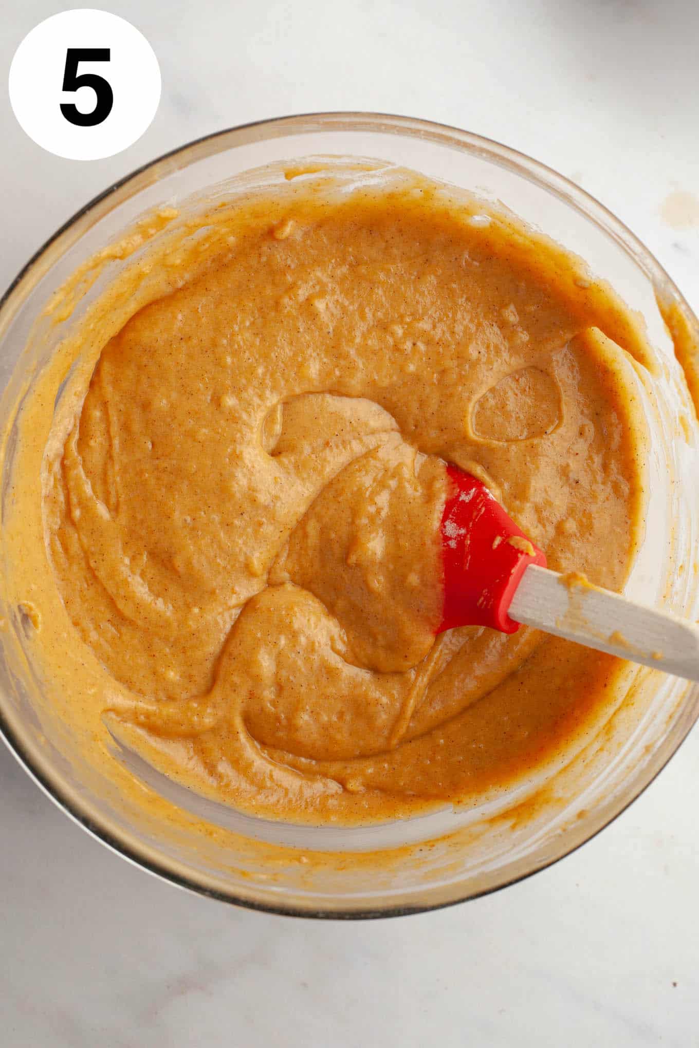 An overhead view of pumpkin cake batter in a glass mixing bowl, with a rubber spatula. 