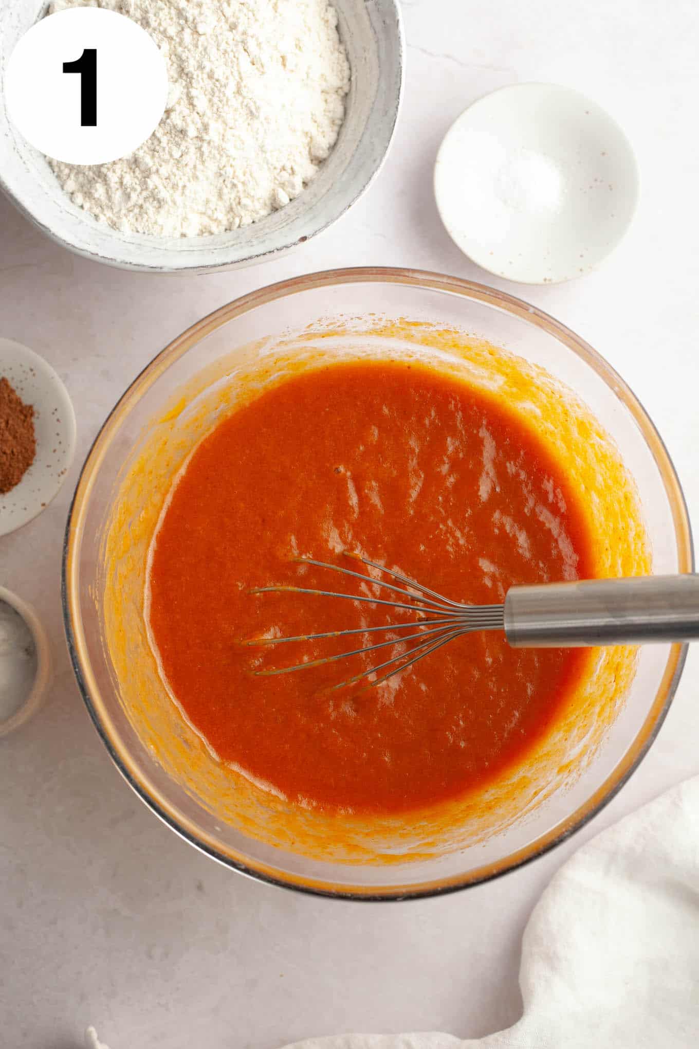 An overhead view of a pumpkin puree mixture in a glass bowl, with a wire whisk. 
