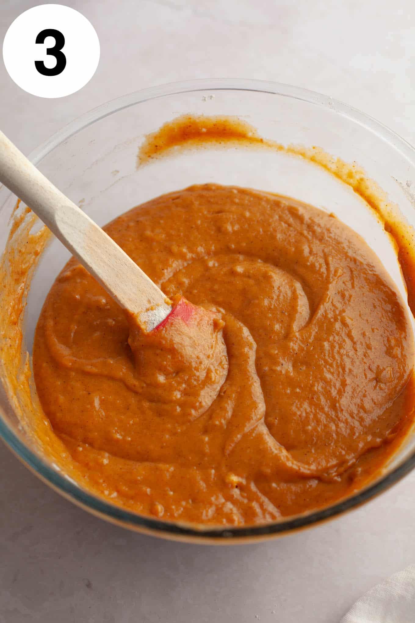 An overhead view of pumpkin bread batter in a glass bowl, with a rubber spatula. 