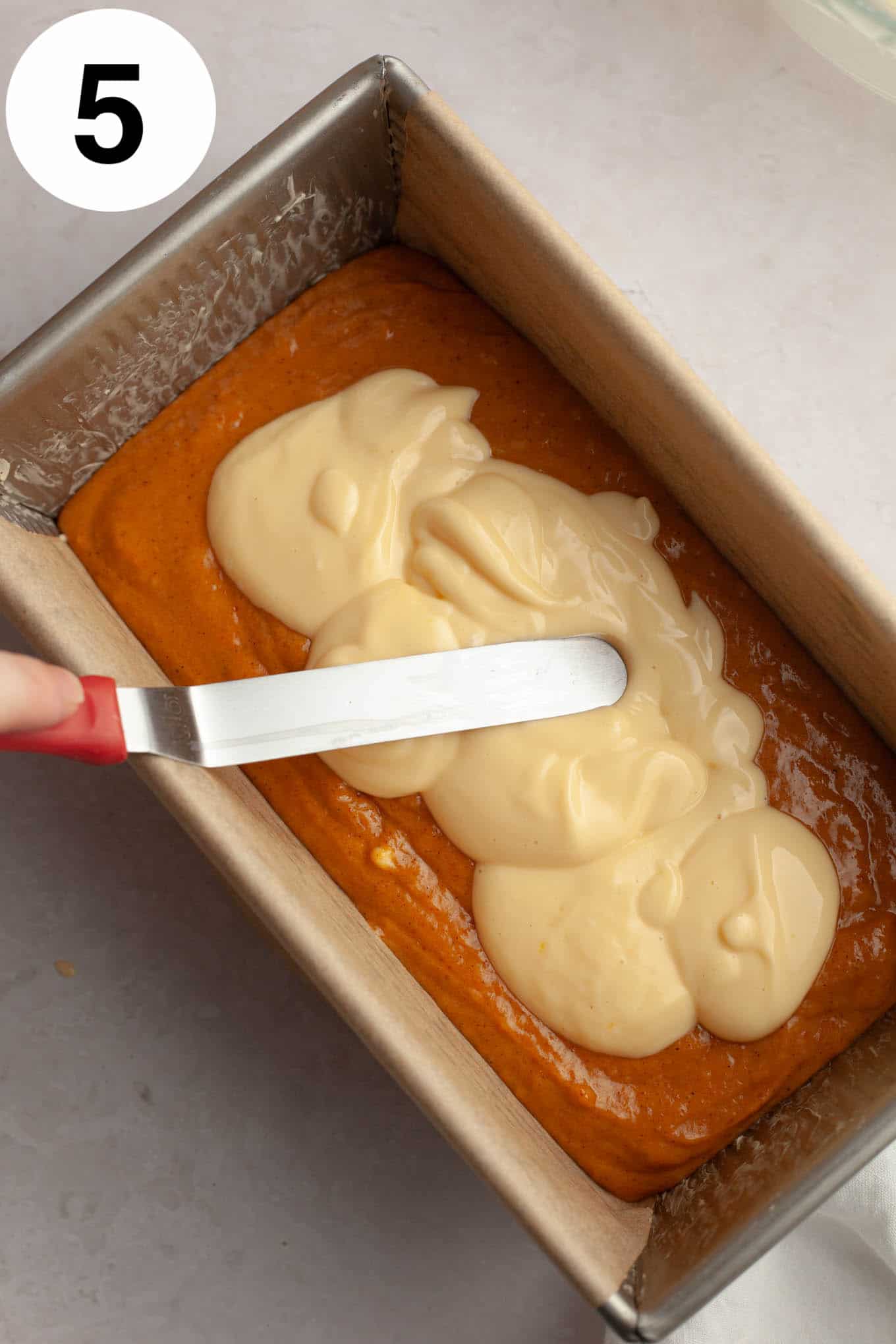 An overhead view of cream cheese filling being spread over pumpkin bread batter in a loaf pan. 