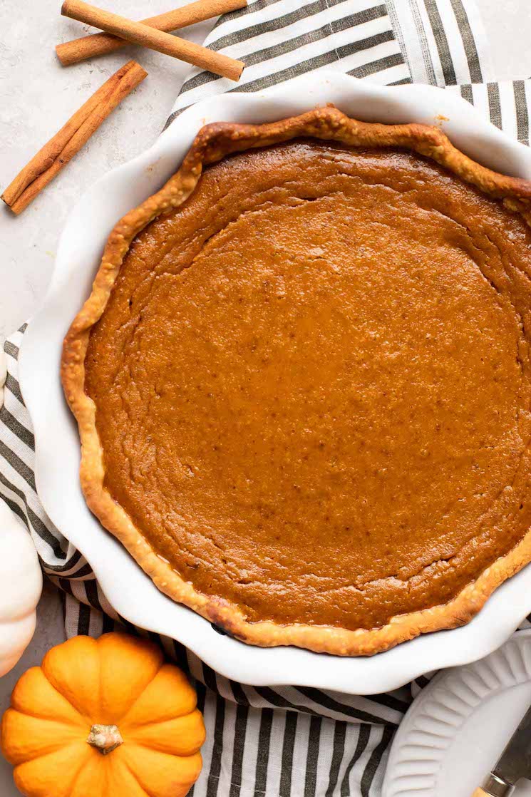 An overhead view of a homemade pumpkin pie in a white pie plate. A mini pumpkin and cinnamon sticks are near it.
