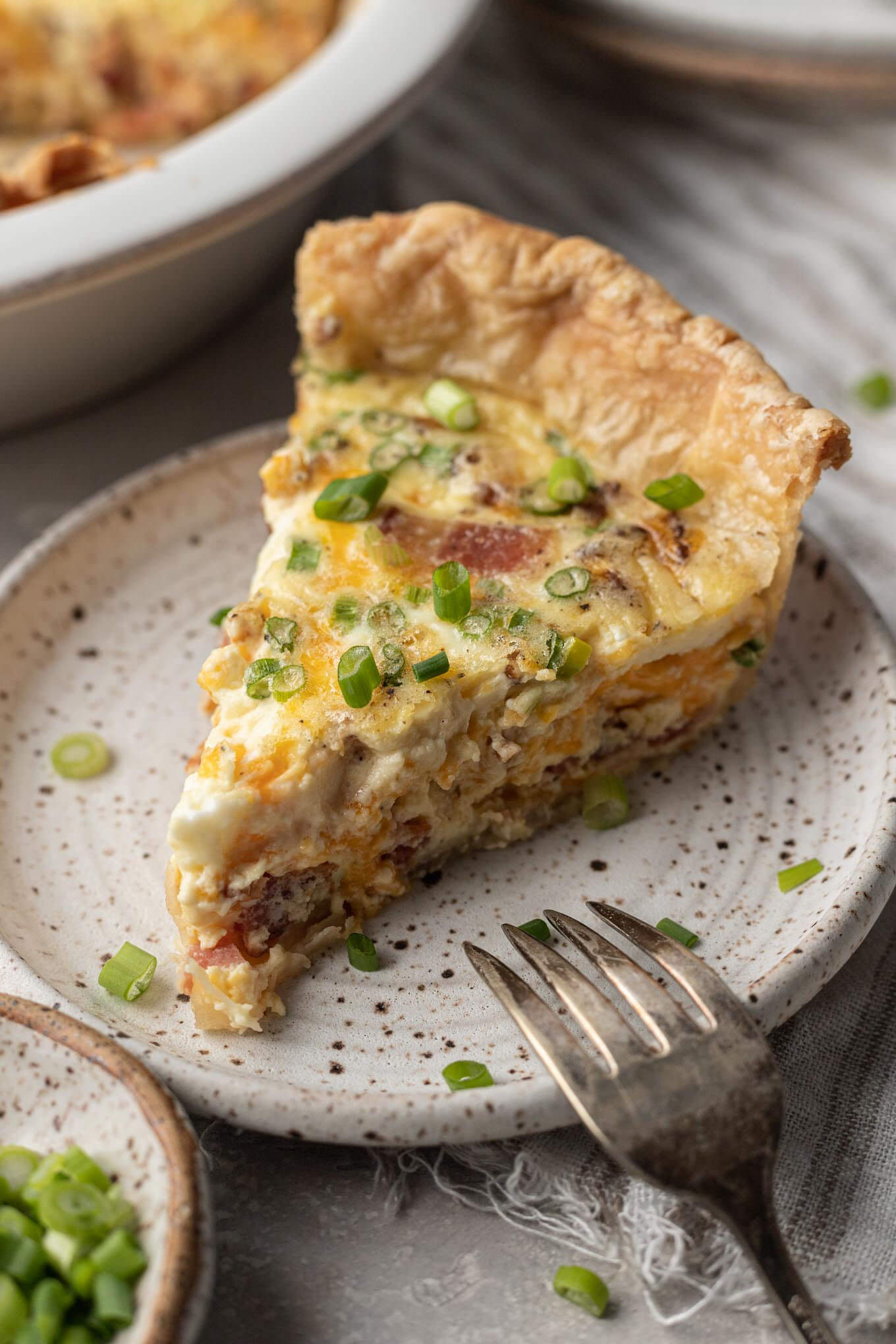 A slice of bacon and cheese quiche on a speckled white plate.