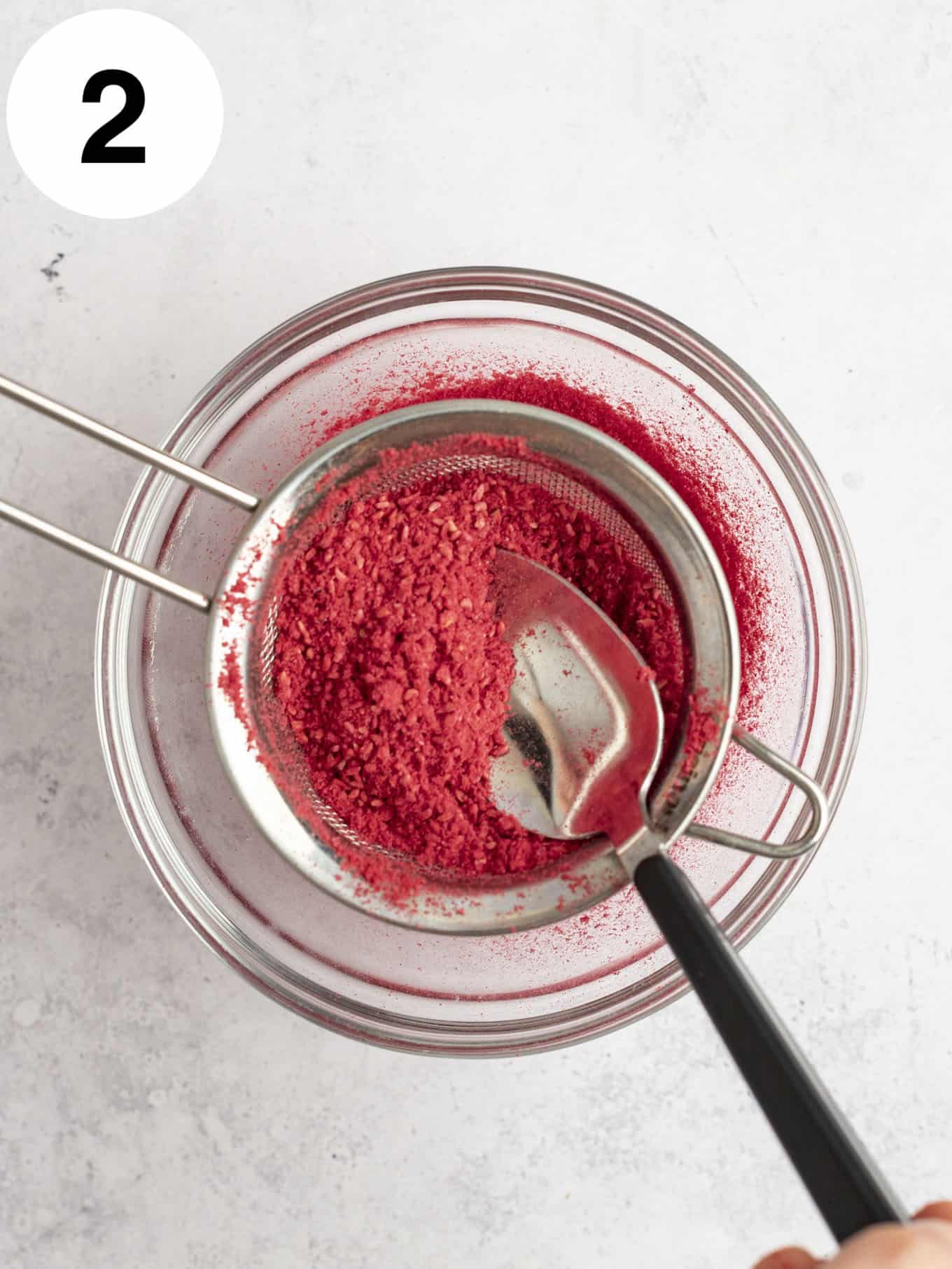 An overhead view of freeze-dried raspberry powder being passed through a mesh sieve. 